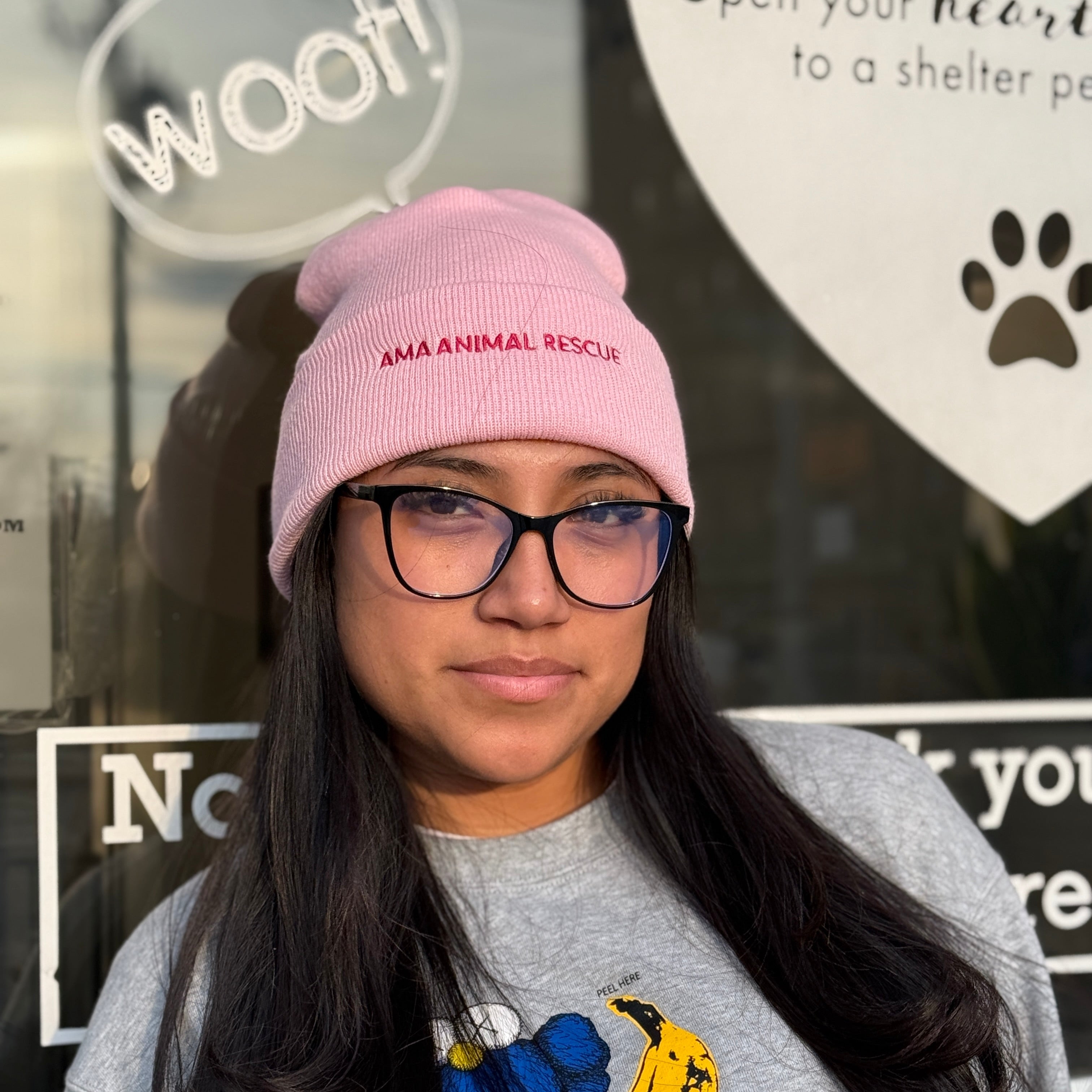 Person wearing a pink beanie with 'AMA Animal Rescue' and a gray sweatshirt with a blue and yellow design, standing in front of AMA Animal rescue in brooklyn