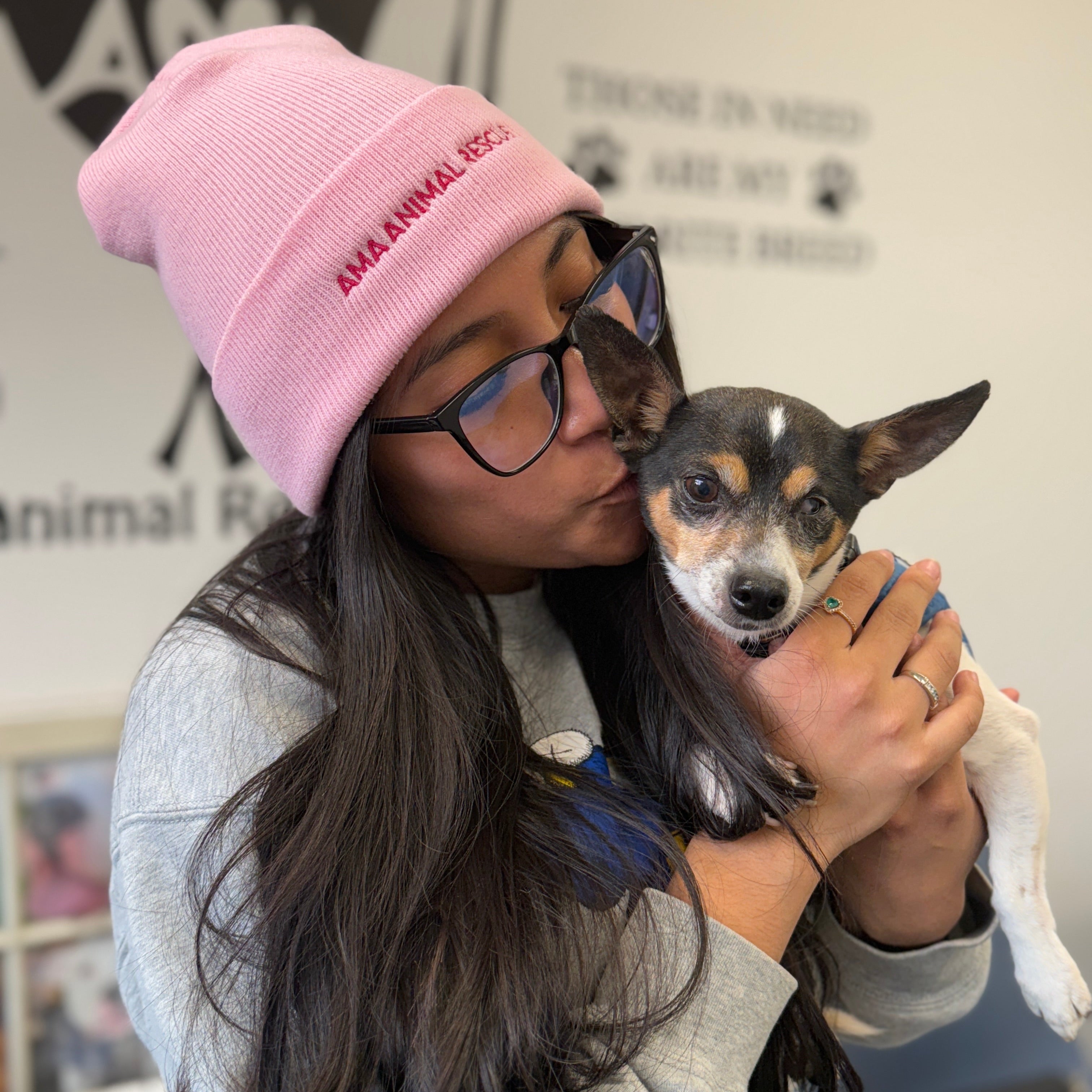 Person wearing a pink beanie with 'AMAAnimal Rescue' holding a small dog indoors.
